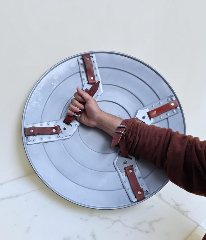 Op de afbeelding: Een zilverkleurig, cirkelvormig schild met een reeks concentrische groeven. Het schild heeft vier bruine leren riemen die met zilveren klinknagels zijn bevestigd. Het schild wordt door een persoon vastgehouden.