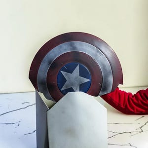 May include: A Captain America shield replica, featuring a red, silver, and blue color scheme. The shield has a central blue star on a blue circle, surrounded by concentric rings of red and silver. The shield is partially resting on a gray stand.