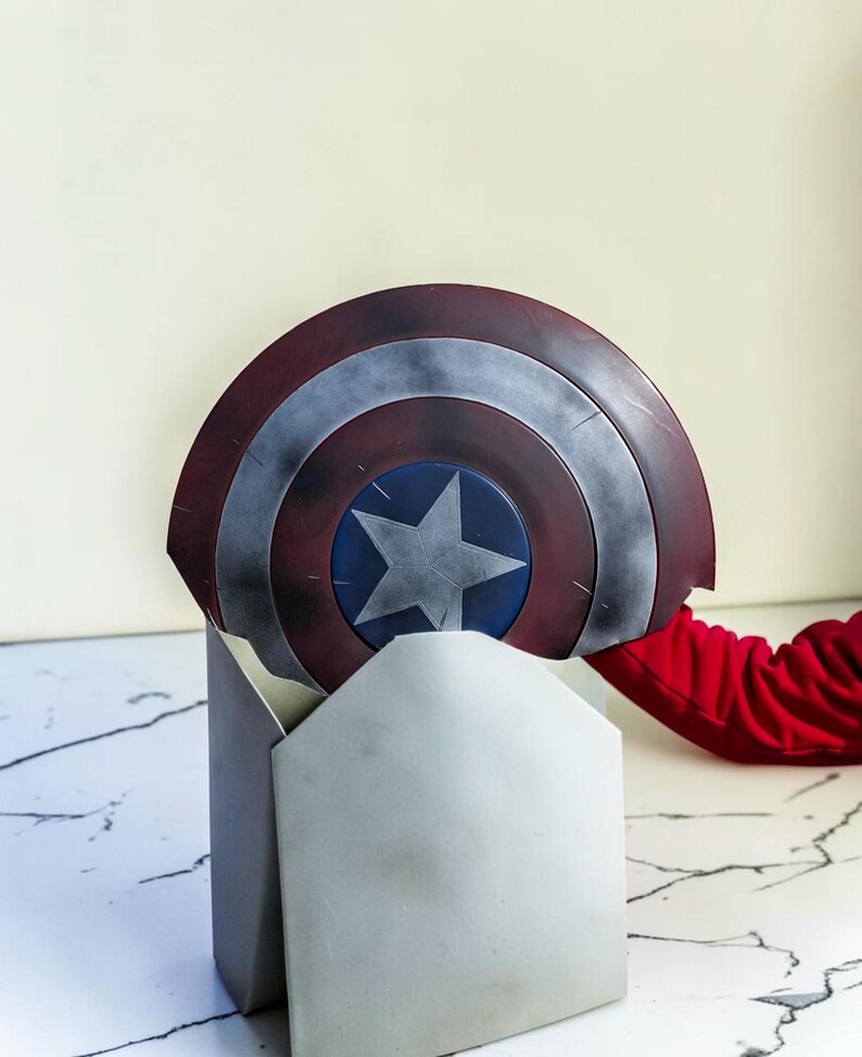 May include: A Captain America shield replica, featuring a red, silver, and blue color scheme. The shield has a central blue star on a blue circle, surrounded by concentric rings of red and silver. The shield is partially resting on a gray stand.