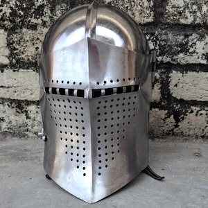 May include: A polished, silver-colored medieval helmet with a rounded top and a pointed visor. The visor features numerous small, evenly spaced holes for ventilation. The helmet is designed to protect the head and face.