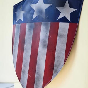May include: A shield with a patriotic design. The top section is blue with three white stars. The lower portion features red and white stripes. The shield has a slightly distressed appearance.