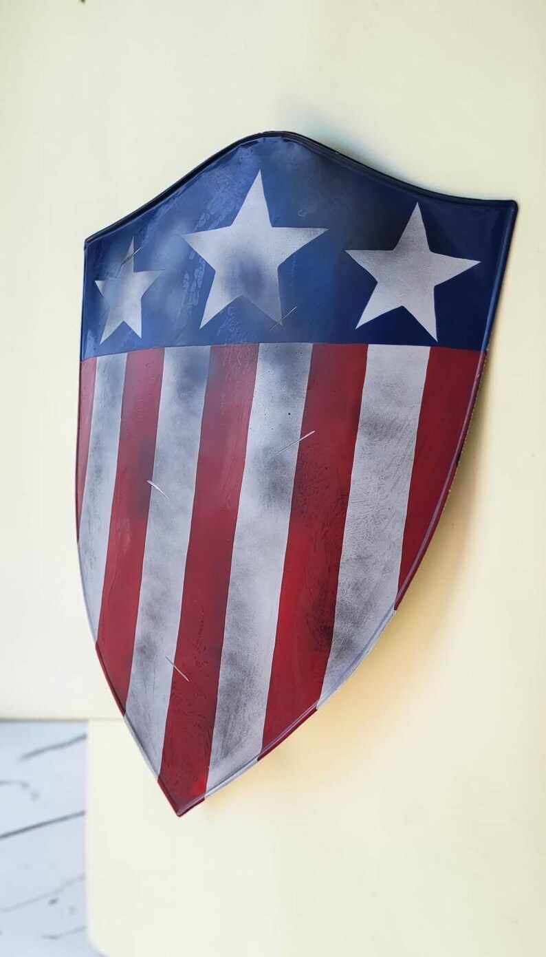 May include: A shield with a patriotic design. The top section is blue with three white stars. The lower portion features red and white stripes. The shield has a slightly distressed appearance.