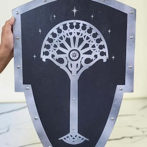 Pode incluir: Um escudo preto com acabamento e rebites prateados. O escudo apresenta um design de árvore prateada estilizado com uma explosão de estrelas no topo e um padrão de estrelas no fundo preto. O escudo é segurado por uma pessoa.