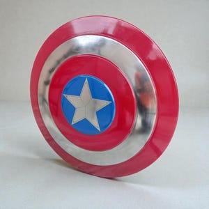 Puede incluir: Un escudo rojo, plateado y azul con un emblema de estrella. El escudo tiene un dise&ntilde;o circular con anillos conc&eacute;ntricos rojos, plateados y un centro azul con una estrella blanca. Un accesorio cl&aacute;sico de superh&eacute;roe.