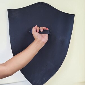 May include: A black shield with a curved top and pointed bottom is held by a hand. The shield has a handle on the back. The shield is against a light-colored wall.