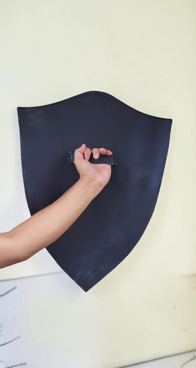 May include: A black shield with a curved top and pointed bottom is held by a hand. The shield has a handle on the back. The shield is against a light-colored wall.