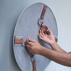 Peut inclure: Un bouclier rond en m&eacute;tal gris avec des sangles en cuir marron et des rivets argent&eacute;s. Le bouclier a un aspect l&eacute;g&egrave;rement vieilli. Les sangles sont fix&eacute;es au bouclier avec des supports m&eacute;talliques. Le bouclier est tenu par une personne.