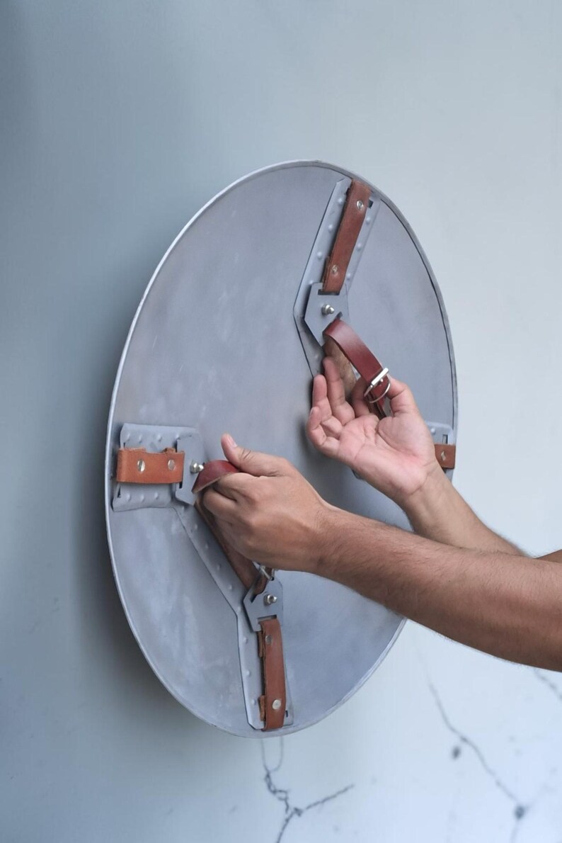 Peut inclure: Un bouclier rond en m&eacute;tal gris avec des sangles en cuir marron et des rivets argent&eacute;s. Le bouclier a un aspect l&eacute;g&egrave;rement vieilli. Les sangles sont fix&eacute;es au bouclier avec des supports m&eacute;talliques. Le bouclier est tenu par une personne.