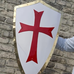 Puede incluir: Un escudo blanco con un diseño de cruz roja y un borde dorado. El escudo se sostiene contra una pared de ladrillos. La cruz tiene extremos puntiagudos. El borde está asegurado con pequeños remaches.