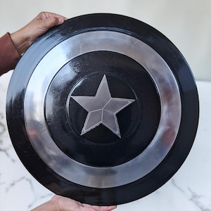 Op de afbeelding: Een zwart en zilver schild met een centraal zilveren sterontwerp. Het schild heeft een cirkelvorm met een zilveren ring rond de centrale ster. Het schild wordt door twee handen vastgehouden.