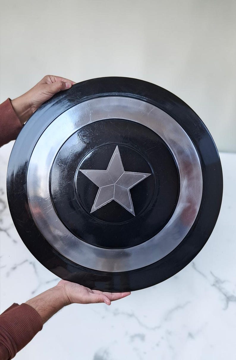 Op de afbeelding: Een zwart en zilver schild met een centraal zilveren sterontwerp. Het schild heeft een cirkelvorm met een zilveren ring rond de centrale ster. Het schild wordt door twee handen vastgehouden.