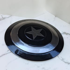 Op de afbeelding: Een zwart en zilver schild met een ster embleem. Het schild is rond met concentrische cirkels en een glanzende afwerking. De centrale ster is donkergrijs.