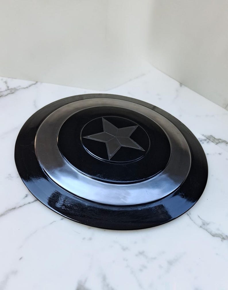 Op de afbeelding: Een zwart en zilver schild met een ster embleem. Het schild is rond met concentrische cirkels en een glanzende afwerking. De centrale ster is donkergrijs.