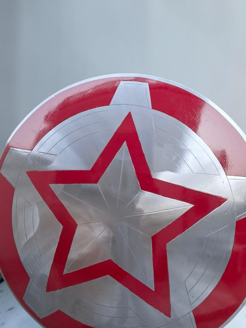 Peut inclure: Un bouclier rond avec un design rouge et argent&eacute;. Le bouclier pr&eacute;sente une grande &eacute;toile rouge au centre, entour&eacute;e de segments argent&eacute;s et d'une bordure rouge. Le bouclier a une finition brillante.