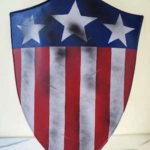 May include: A shield with a patriotic design, featuring a blue top with white stars and red and white stripes. The shield has a curved shape and a distressed, aged appearance.