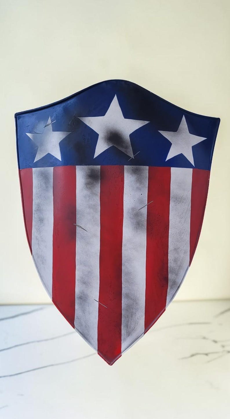 May include: A shield with a patriotic design, featuring a blue top with white stars and red and white stripes. The shield has a curved shape and a distressed, aged appearance.