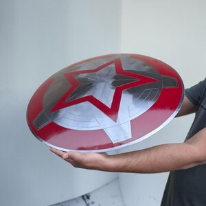 Peut inclure: Un bouclier rond m&eacute;tallique avec un bord rouge et une grande &eacute;toile argent&eacute;e au centre. Le bouclier a une finition brillante. Il s'agit d'un accessoire ou d'un objet de collection.