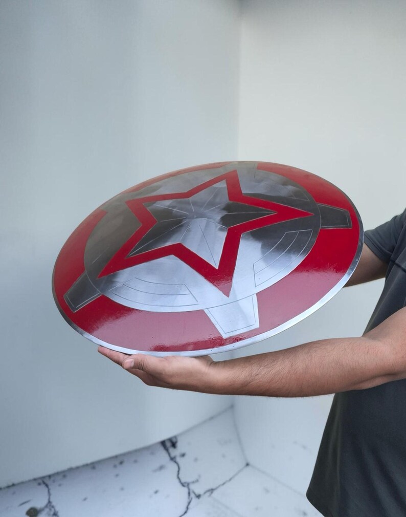 Peut inclure: Un bouclier rond m&eacute;tallique avec un bord rouge et une grande &eacute;toile argent&eacute;e au centre. Le bouclier a une finition brillante. Il s'agit d'un accessoire ou d'un objet de collection.
