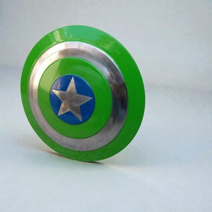 Pu&ograve; includere: Uno scudo verde e argento con un cerchio centrale blu e una stella argentata. Lo scudo ha una finitura lucida e un design circolare, che ricorda l'emblema di un supereroe.