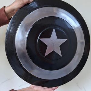 Op de afbeelding: Een zwart en zilver schild met een centraal zilveren sterontwerp. Het schild heeft een cirkelvorm met concentrische ringen van zwart en zilver. Het schild wordt door twee handen vastgehouden.