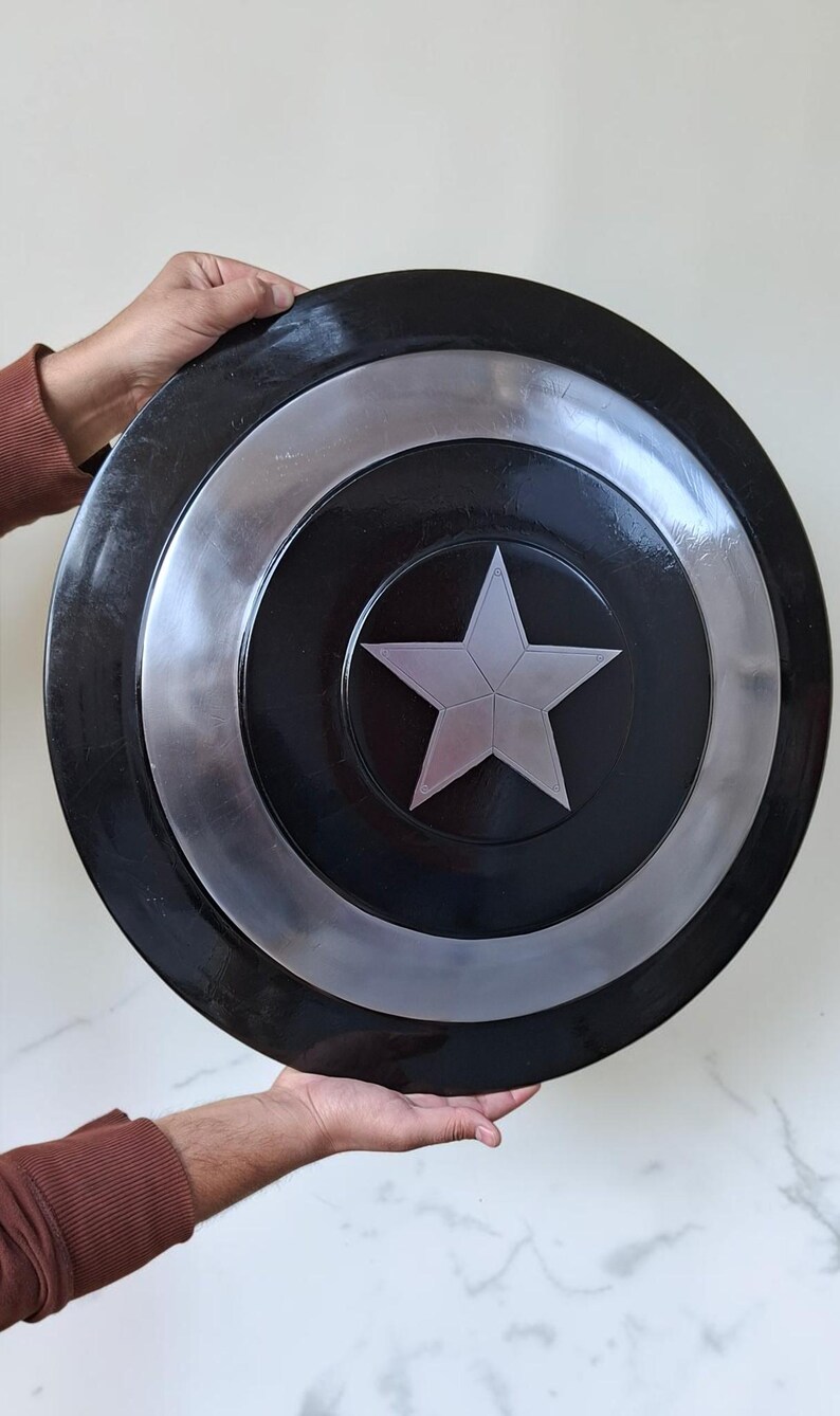 Op de afbeelding: Een zwart en zilver schild met een centraal zilveren sterontwerp. Het schild heeft een cirkelvorm met concentrische ringen van zwart en zilver. Het schild wordt door twee handen vastgehouden.
