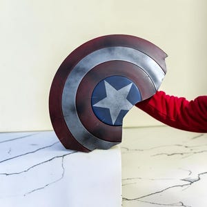May include: A Captain America shield, featuring a red, white, and blue color scheme. The shield has a star in the center and a distressed, battle-worn appearance. The shield is held by a person wearing a red long-sleeve shirt.