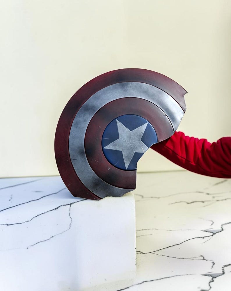 May include: A Captain America shield, featuring a red, white, and blue color scheme. The shield has a star in the center and a distressed, battle-worn appearance. The shield is held by a person wearing a red long-sleeve shirt.