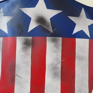May include: A shield-shaped object with a patriotic design. The top section is blue with white stars, and the lower section features red and white stripes. The design has a distressed, aged appearance.