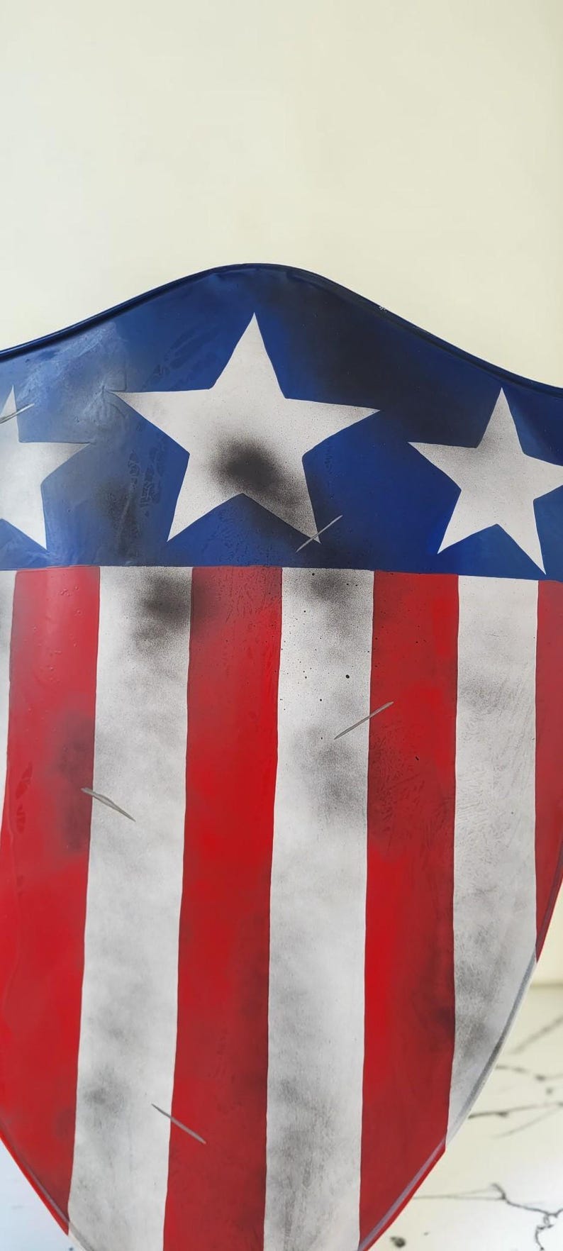 May include: A shield-shaped object with a patriotic design. The top section is blue with white stars, and the lower section features red and white stripes. The design has a distressed, aged appearance.