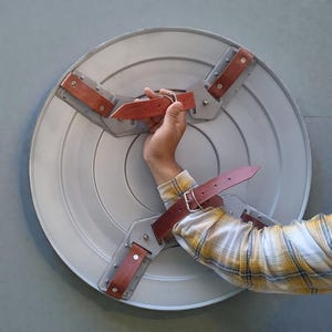 Puede incluir: Un escudo circular gris con correas y asa de cuero marr&oacute;n. El escudo tiene una apariencia met&aacute;lica y est&aacute; dise&ntilde;ado con una serie de c&iacute;rculos conc&eacute;ntricos. Las correas de cuero est&aacute;n remachadas al marco del escudo.