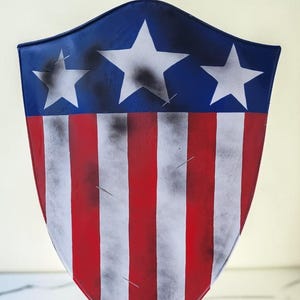 May include: A shield with a blue top section featuring three white stars. The lower portion has red and white vertical stripes. The shield has a distressed, weathered appearance, with black accents and a slightly curved shape.