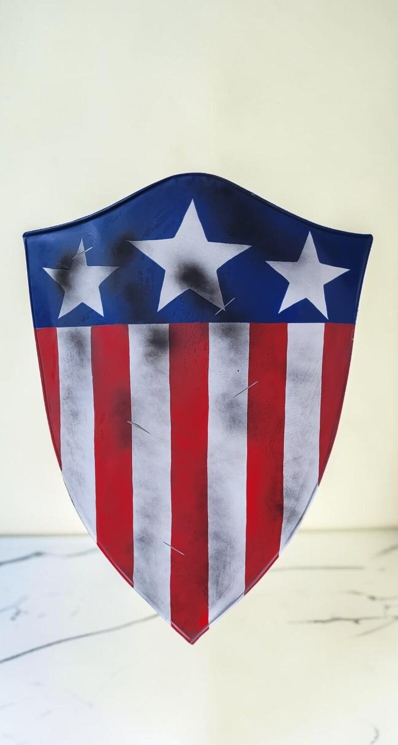 May include: A shield with a blue top section featuring three white stars. The lower portion has red and white vertical stripes. The shield has a distressed, weathered appearance, with black accents and a slightly curved shape.