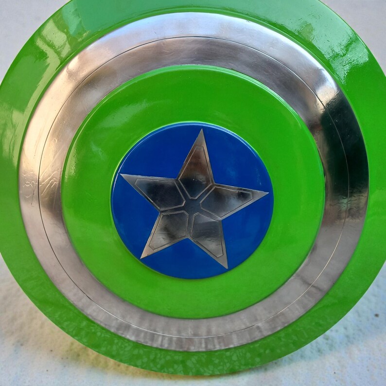 Pu&ograve; includere: Uno scudo verde e argento con un centro blu e una stella argentata. Lo scudo presenta un design circolare con anelli concentrici verdi, argento e blu. La stella &egrave; un design a cinque punte.