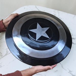 Op de afbeelding: Een zwart en zilver schild met een zilveren ster in het midden. Het schild heeft een cirkelvorm met concentrische ringen van zwart en zilver. Het schild wordt door twee handen vastgehouden.