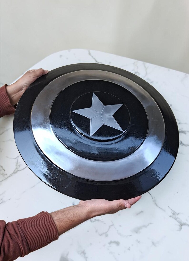 Op de afbeelding: Een zwart en zilver schild met een zilveren ster in het midden. Het schild heeft een cirkelvorm met concentrische ringen van zwart en zilver. Het schild wordt door twee handen vastgehouden.