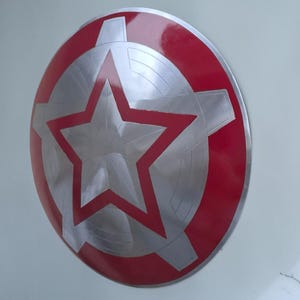 Peut inclure: Un bouclier rond avec un bord rouge et un centre argent&eacute;. Le centre pr&eacute;sente une grande &eacute;toile rouge et un motif argent&eacute; en forme de roue dent&eacute;e. Le bouclier est probablement en m&eacute;tal et a une finition brillante.