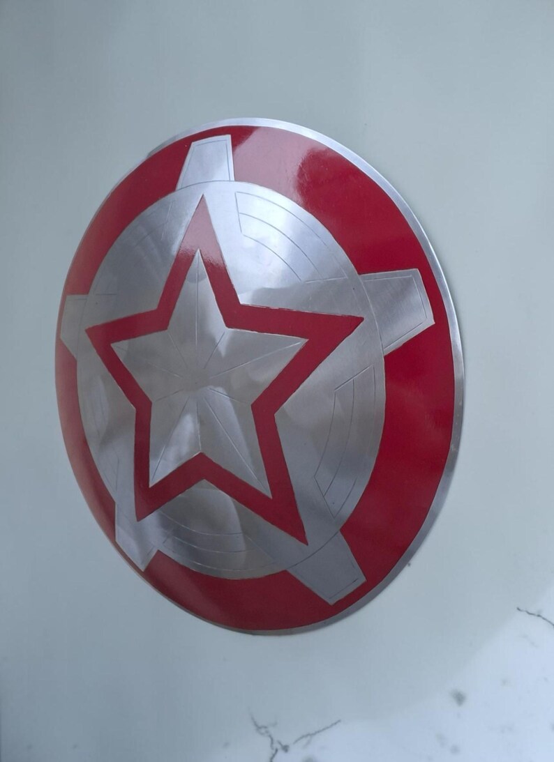 Peut inclure: Un bouclier rond avec un bord rouge et un centre argent&eacute;. Le centre pr&eacute;sente une grande &eacute;toile rouge et un motif argent&eacute; en forme de roue dent&eacute;e. Le bouclier est probablement en m&eacute;tal et a une finition brillante.
