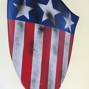 May include: A shield with a patriotic design, featuring a blue top with three white stars and red and white stripes. The shield has a distressed, weathered appearance, with black smudges and scratches.