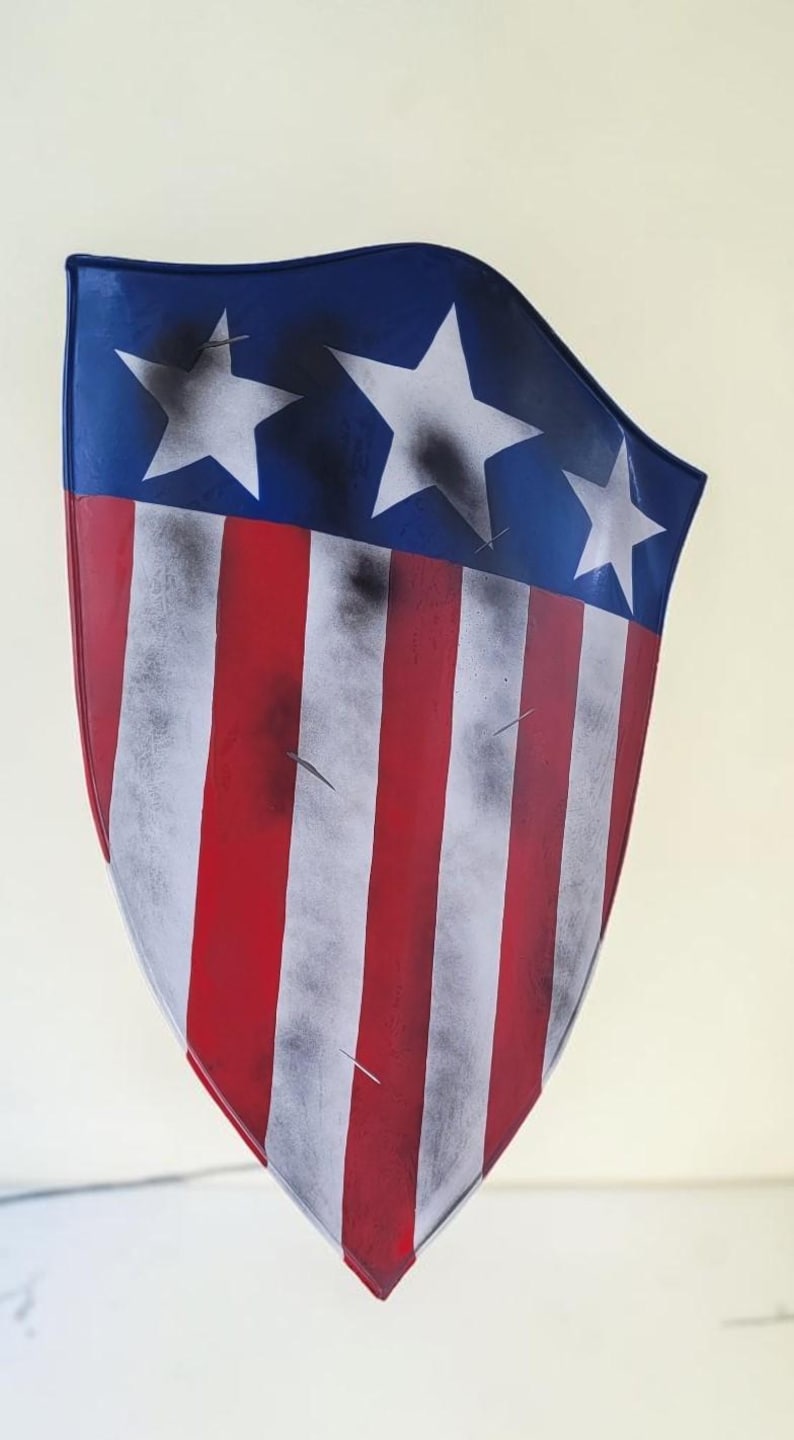 May include: A shield with a patriotic design, featuring a blue top with three white stars and red and white stripes. The shield has a distressed, weathered appearance, with black smudges and scratches.