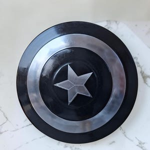 Op de afbeelding: Een zwart en zilver schild met een centraal zilveren sterembleem. Het schild heeft concentrische cirkelvormige banden van zwart en zilver. De ster is een vijfpuntig ontwerp. Het schild is een rekwisiet of verzamelobject.