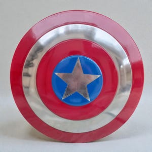 Puede incluir: Un escudo redondo con un borde exterior rojo, un anillo plateado en el medio y un centro azul. Una estrella plateada est&aacute; en el centro del c&iacute;rculo azul. El escudo es una r&eacute;plica del escudo de un superh&eacute;roe.