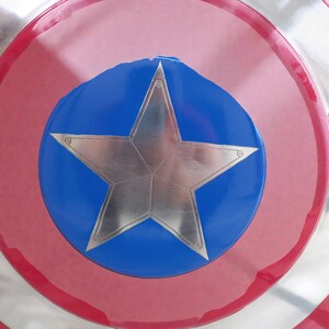 Puede incluir: Primer plano de un escudo con una estrella plateada sobre un c&iacute;rculo azul, sobre un fondo rojo y plateado. El escudo tiene un dise&ntilde;o circular con un anillo exterior rojo y un anillo interior plateado.