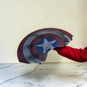 May include: A broken Captain America shield, featuring a red, white, and blue color scheme. The shield has a central blue circle with a white star, surrounded by red and white concentric rings. The shield is held by a person wearing a red long-sleeve shirt.