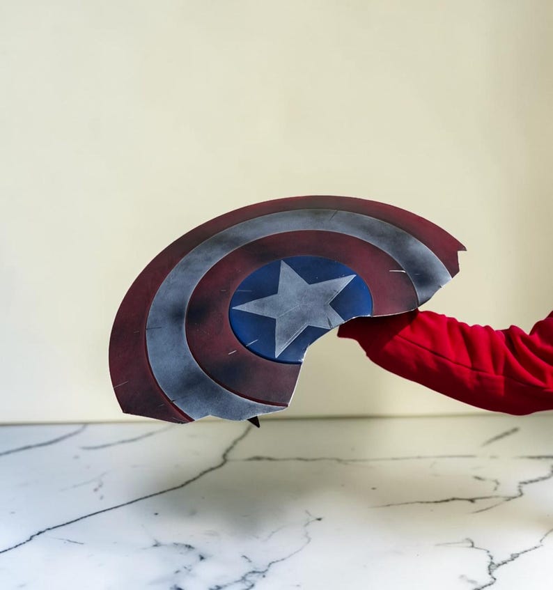 May include: A broken Captain America shield, featuring a red, white, and blue color scheme. The shield has a central blue circle with a white star, surrounded by red and white concentric rings. The shield is held by a person wearing a red long-sleeve shirt.