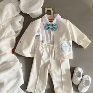 Könnte beinhalten: Ein weißes Baby-Outfit-Set, bestehend aus Jacke, Hose, Hemd, Fliege, Hut und Schuhen. Die Jacke hat ein Kreuz auf der Tasche gestickt. Die Fliege ist hellblau. Das Outfit wird auf einem Holzkleiderbügel präsentiert.