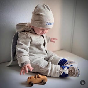 Puede incluir: Un bebé con un mono gris con capucha y orejas de conejo, gorro a juego con "Samir" bordado en azul y zapatillas de crochet con detalles azules. Un pequeño coche de juguete de madera está frente al bebé.