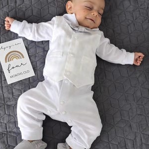Könnte beinhalten: Ein Baby in einem weißen Outfit, bestehend aus einem Hemd mit Knöpfen, einer Weste und einer Hose, mit einer grauen Fliege und passenden Babyschuhen. Eine Karte neben dem Baby zeigt "four months old" mit einer Regenbogen-Grafik. Das Baby liegt auf einer grauen Steppdecke.