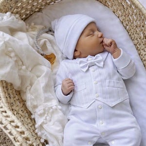 Könnte beinhalten: Ein schlafendes Baby in einem weißen Outfit mit Fliege und passendem Hut, liegend in einem geflochtenen Korb. Das Outfit hat ein Design mit Knopfleiste. Eine weiße Decke mit Pompon-Besatz liegt im Korb.