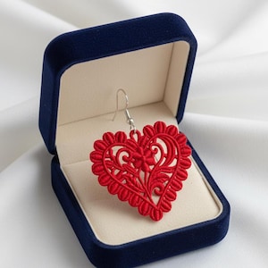 May include: A red heart-shaped earring with intricate floral lace design, displayed in a dark blue velvet jewelry box. The earring has a silver hook and is set against a white background.