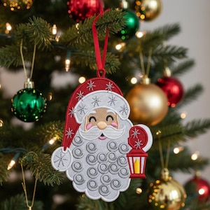 May include: Embroidered Santa Claus Christmas ornament with a red hat and a white beard, holding a lantern. The ornament is hanging on a Christmas tree with other ornaments.